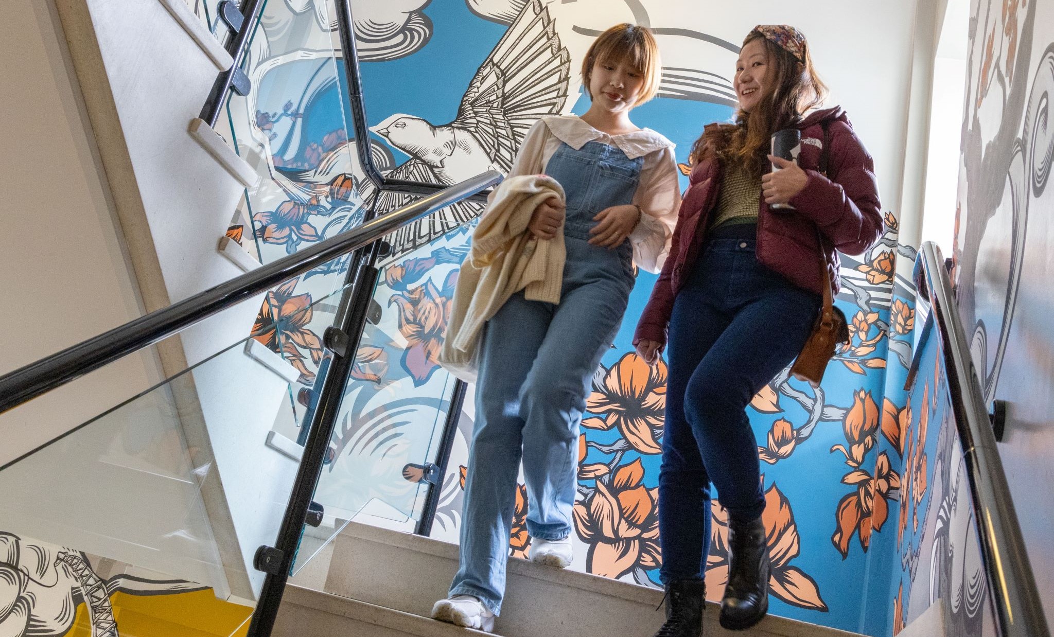 Two female students descend the stairs in the School of Education. behind them is a mural on the wall 